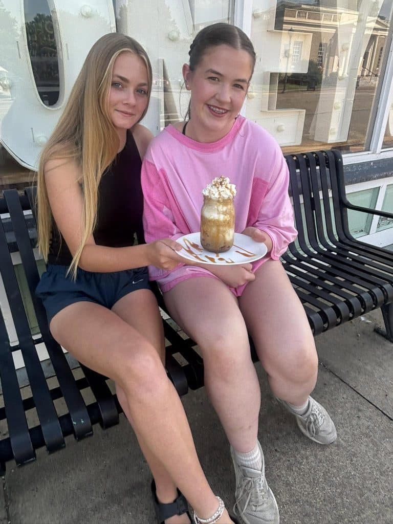 Two teenage girls sit on a black metal bench outside a storefront, sharing a caramel frappé with whipped cream on a white plate.